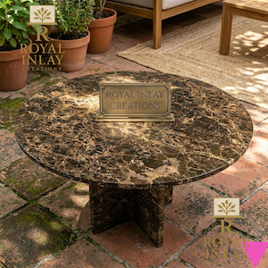 Op de afbeelding: Een ronde salontafel met een bruin, marmerachtig oppervlak. De tafel heeft een donkerbruin onderstel met een uniek ontwerp. Een klein bordje met de tekst "ROYAL INLAY CREATIONS" staat op de tafel. De tafel staat buiten.