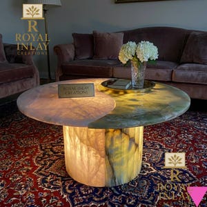 May include: A round coffee table with a two-tone onyx top and a cylindrical base. The top is divided into pink and green sections. The base and the pink section of the top are illuminated. The table is on a patterned red rug. The text "Royal Inlay Creations" is visible.