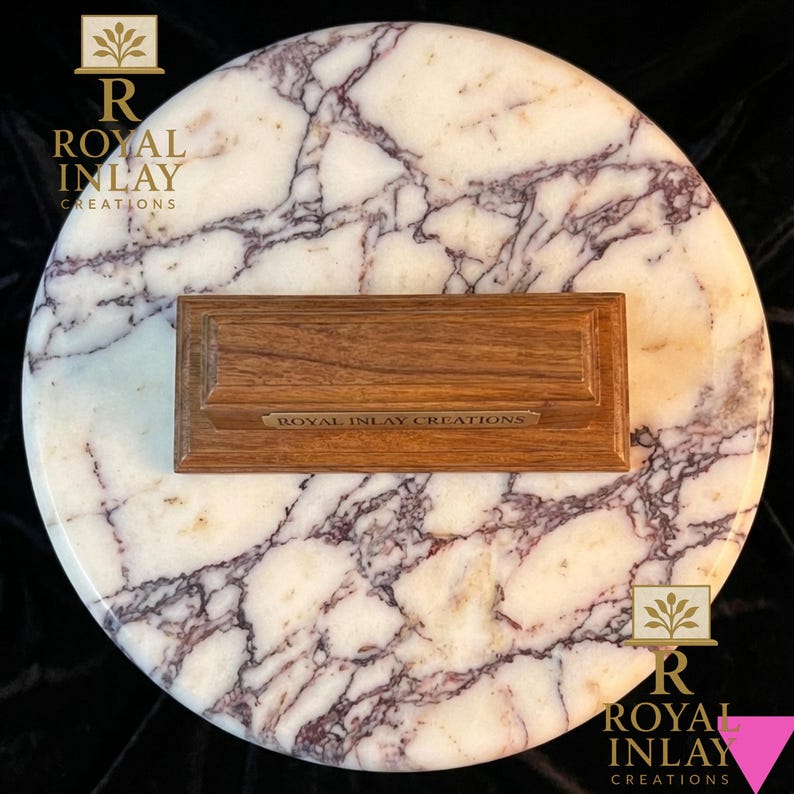 May include: A rectangular wooden block with a stepped base and a small metal plaque reading ROYAL INLAY CREATIONS, centred on a circular white marble slab featuring prominent dark purple veining throughout the stone surface.