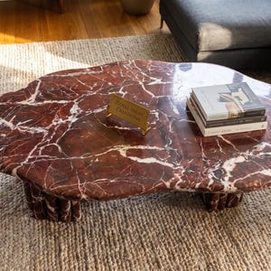 Puede incluir: Una mesa de centro con un tablero de forma orgánica irregular hecho de mármol rojo intenso con vetas blancas prominentes. La mesa se apoya sobre varias patas cilíndricas de mármol e incluye un pequeño letrero dorado Royal Inlay Creations.