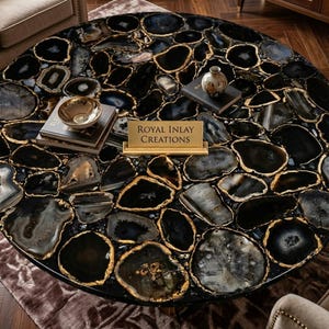 May include: A round coffee table with a black and gray agate stone top, featuring gold-colored edging. The table is adorned with decorative items, including a small gold bowl, books, and a vase. The table has the text "Royal Inlay Creations".