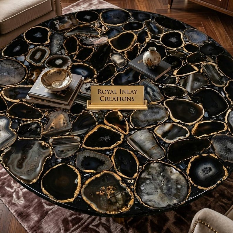 May include: A round coffee table with a black and gray agate stone top, featuring gold-colored edging. The table is adorned with decorative items, including a small gold bowl, books, and a vase. The table has the text "Royal Inlay Creations".
