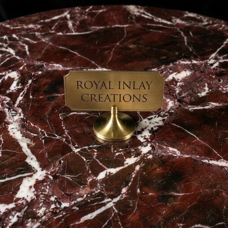 Puede incluir: Un portacarteles de lat&oacute;n con el texto "ROYAL INLAY CREATIONS" sobre una superficie de m&aacute;rmol rojo con vetas blancas. El portacarteles tiene una base redonda y un soporte curvo.