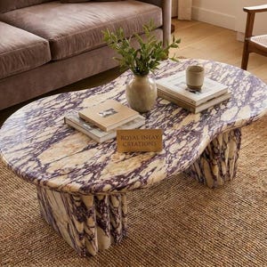 Puede incluir: Una mesa de centro ovalada de mármol veteado en morado y crema con base estriada. La superficie sostiene un jarrón de cerámica con follaje verde, dos pilas de libros, una taza pequeña y una placa dorada con ROYAL INLAY CREATIONS.