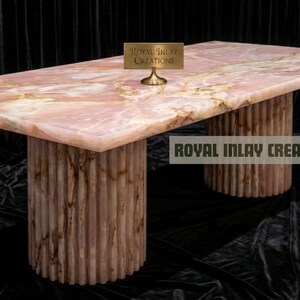 May include: A rectangular pink onyx table with two cylindrical bases. The tabletop has a marbled appearance with light and dark pink hues. The bases are vertically ribbed. The text "Royal Inlay Creations" is visible.