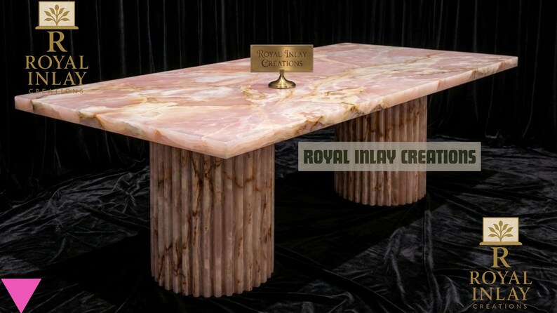 May include: A rectangular pink onyx table with two cylindrical bases. The tabletop has a marbled appearance with light and dark pink hues. The bases are vertically ribbed. The text "Royal Inlay Creations" is visible.