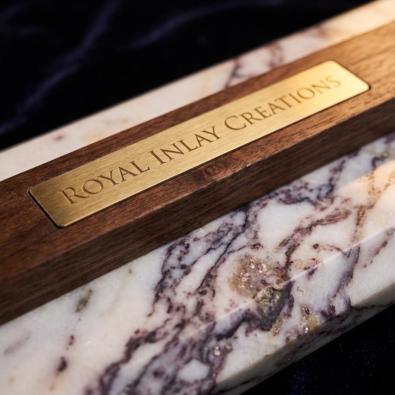 May include: A rectangular piece of white marble with dark purple veining paired with a dark brown wooden block. A rectangular brass-coloured metal plate is inlaid into the wood, featuring the engraved text ROYAL INLAY CREATIONS in capital letters.
