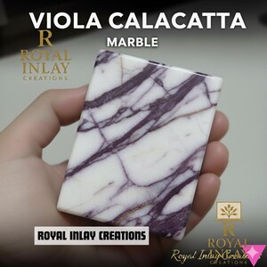 May include: A rectangular piece of Viola Calacatta marble featuring a white base with prominent deep purple veining. The text VIOLA CALACATTA MARBLE and ROYAL INLAY CREATIONS is visible on the surface. The stone has a smooth, polished finish.