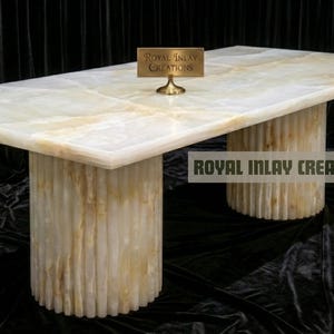 Puede incluir: Mesa rectangular de ónix con superficie veteada en crema claro y dorado. La mesa descansa sobre dos bases cilíndricas con detalles acanalados verticales. Un pequeño letrero dice "Royal Inlay Creations". El fondo es negro.