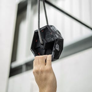 May include: A black hexagonal climbing hold with red numbers hanging from a rope. A hand reaches up to grab the hold.