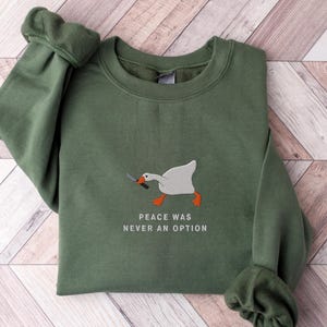 以下が含まれることがあります： ナイフを持った白いガチョウと「Peace was never an option」という文字が書かれた緑色のスウェットシャツ。