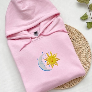 Puede incluir: Una sudadera con capucha rosa con un diseño de sol y luna bordado en la parte delantera. El sol es amarillo con una cara y la luna es gris con una cara dormida.