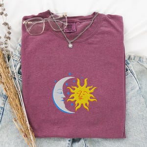 May include: A maroon t-shirt featuring an embroidered design of a yellow sun and a blue crescent moon with a face. The sun and moon are overlapping.