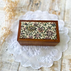May include: A small, rectangular wooden box with a floral tapestry lid. The tapestry features a pattern of green, pink, and cream-colored flowers. The box is set on a white lace doily, creating a vintage aesthetic.