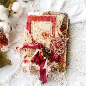 Puede incluir: Un diario rectangular con cubierta de tela que presenta capas de textiles rojos y beige, bordados intrincados, encaje y adornos florales con lentejuelas. La cubierta está atada con cintas rojas deshilachadas con dijes y cuentas.