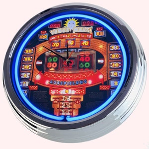May include: A round, silver-framed clock with a blue neon ring around the edge. The clock face features a colourful image of a slot machine with the words "VENUS MULTI" and "AUSSPIELUNG".