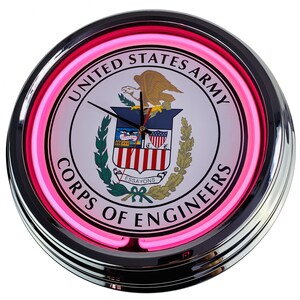 May include: A pink neon clock with a white face featuring the United States Army Corps of Engineers seal. The seal depicts a bald eagle with a shield and the words "United States Army Corps of Engineers" in a circle around the seal.