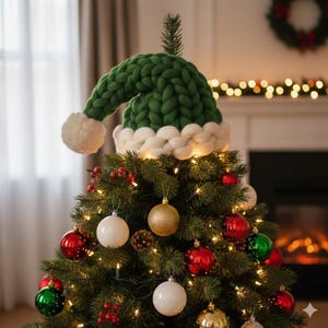 May include: A Christmas tree decorated with ornaments and lights, topped with a green and white knitted Santa hat. The tree is adorned with red, gold, and white ornaments, and a string of lights. A cozy holiday scene.