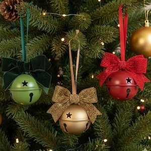 Puede incluir: Tres adornos navideños de campana. Una campana verde con un lazo verde oscuro, una campana dorada con un lazo dorado y una campana roja con un lazo rojo brillante. Cada campana tiene un recorte de estrella y un diseño de ojo de cerradura.