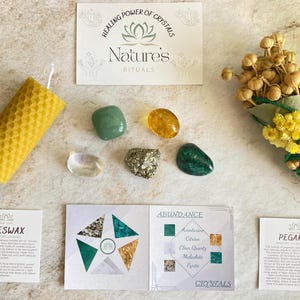 May include: A green and gold crystal kit for abundance rituals. The kit includes a beeswax candle, a bundle of dried flowers, and five crystals: aventurine, citrine, clear quartz, malachite, and pyrite. The kit also includes two cards with information about the crystals and their uses.