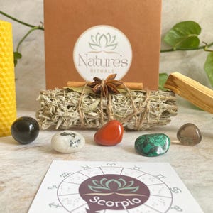 May include: A Scorpio zodiac kit with a white sage smudge stick, a palo santo stick, a honeycomb candle, a red stone, a black stone, a green stone, a brown stone, and a zodiac wheel with the word "Scorpio" printed on it.
