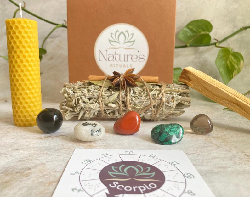 May include: A Scorpio zodiac kit with a white sage smudge stick, a palo santo stick, a honeycomb candle, a red stone, a black stone, a green stone, a brown stone, and a zodiac wheel with the word "Scorpio" printed on it.