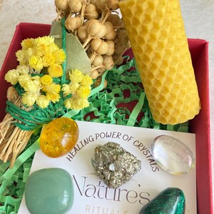 May include: A red box filled with green tissue paper contains a yellow honeycomb candle, a bundle of dried flowers, a green aventurine crystal, a pyrite crystal, a clear quartz crystal, and an amber stone. The box also includes a card with the text "Healing Power of Crystals Nature's Rituals".