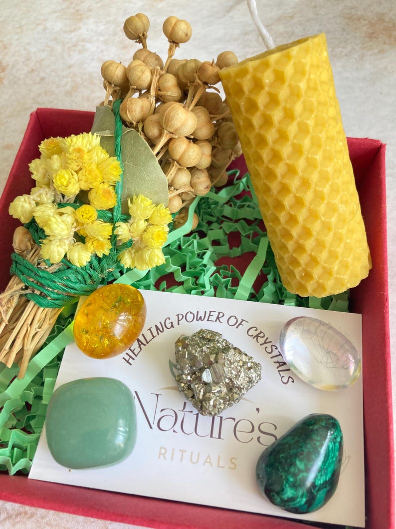 May include: A red box filled with green tissue paper contains a yellow honeycomb candle, a bundle of dried flowers, a green aventurine crystal, a pyrite crystal, a clear quartz crystal, and an amber stone. The box also includes a card with the text "Healing Power of Crystals Nature's Rituals".