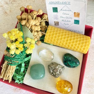 May include: A red box filled with a variety of items for an abundance ritual. The box contains a bundle of dried flowers, a honeycomb candle, a clear quartz crystal, a green aventurine crystal, a malachite crystal, a pyrite crystal, and a citrine crystal. The box also includes a card with the words "Abundance" and a list of crystals.