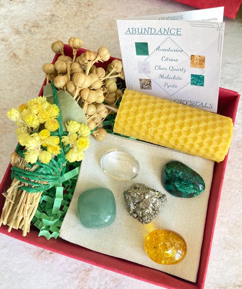 May include: A red box filled with a variety of items for an abundance ritual. The box contains a bundle of dried flowers, a honeycomb candle, a clear quartz crystal, a green aventurine crystal, a malachite crystal, a pyrite crystal, and a citrine crystal. The box also includes a card with the words "Abundance" and a list of crystals.