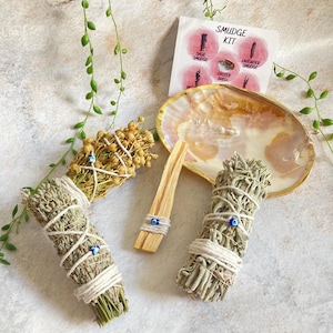 May include: A smudge kit with three bundles of dried herbs, a palo santo stick, and a decorative seashell. The bundles are tied with twine and have blue evil eye charms. The card reads "Smudge Kit" with the names of the herbs: Sage Smudge, Oyster Shell, Lavender Smudge.