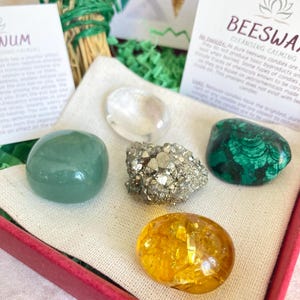 May include: A set of five crystals in a red box with a white cloth lining. The crystals are green, clear, gold, brown, and green. The box also includes a small card with the text "Beeswax Candles Cleansing Calming" and a lotus flower symbol.