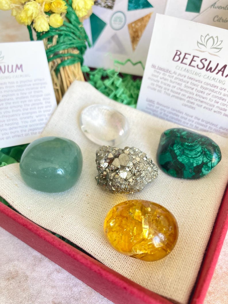 May include: A set of five crystals in a red box with a white cloth lining. The crystals are green, clear, gold, brown, and green. The box also includes a small card with the text "Beeswax Candles Cleansing Calming" and a lotus flower symbol.