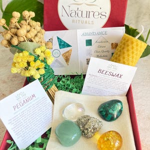 May include: A red box filled with green and yellow crystals, a small bundle of dried flowers, a beeswax candle, and a small white pouch. The box is labelled "Nature's Rituals" and "Abundance". The crystals are labelled "Aventurine, Citrine, Clear Quartz, Malachite". The candle is labelled "Beeswax Cleansing Calming".