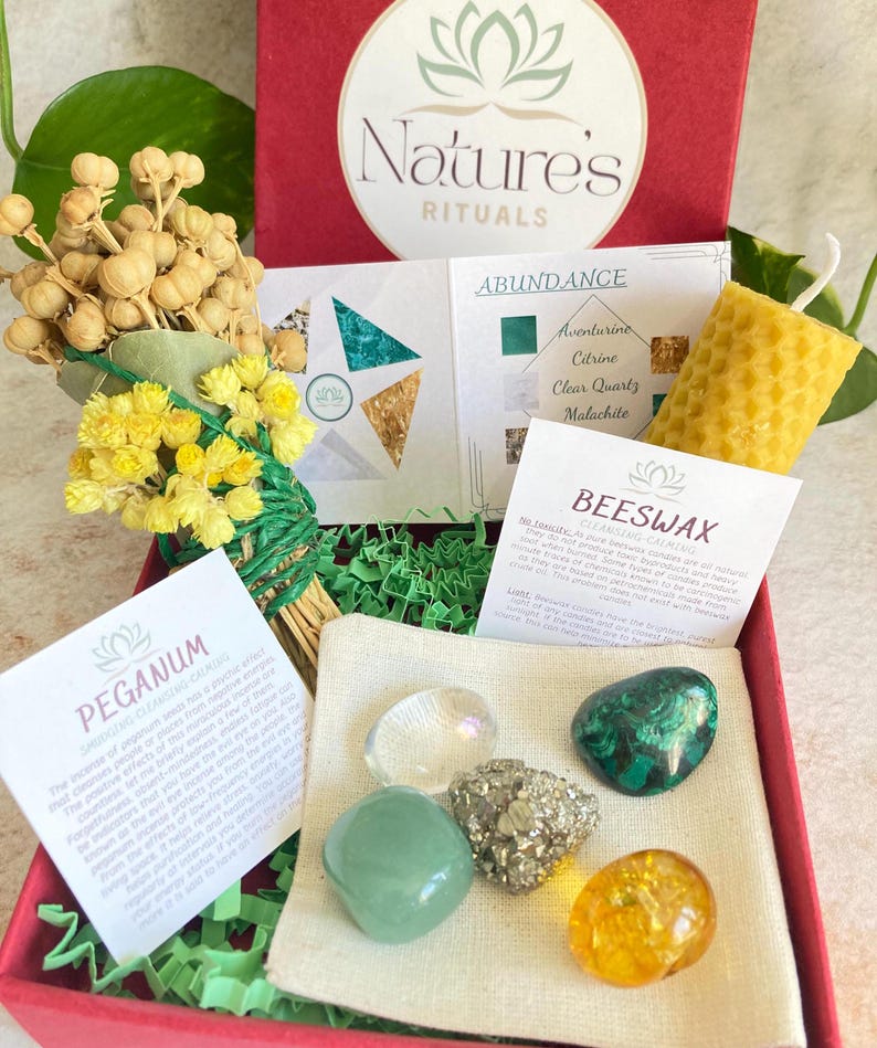 May include: A red box filled with green and yellow crystals, a small bundle of dried flowers, a beeswax candle, and a small white pouch. The box is labelled "Nature's Rituals" and "Abundance". The crystals are labelled "Aventurine, Citrine, Clear Quartz, Malachite". The candle is labelled "Beeswax Cleansing Calming".
