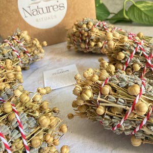 May include: Four bundles of dried herbs tied with red and white string. The bundles are made of dried lavender and small, round, beige seeds. The bundles are arranged on a light beige surface. The text "Nature's Rituals" is visible on a brown box in the background.