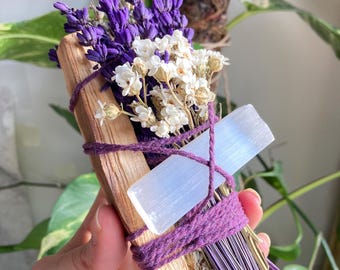 May include: A purple and white dried flower smudge stick with a clear selenite crystal. The smudge stick is tied with purple twine and is held in a hand.