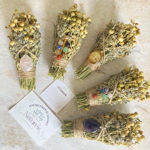 May include: Five bundles of dried herbs tied with twine and adorned with crystals. Each bundle features a different crystal or set of crystals, including amethyst, rose quartz, clear quartz, and a set of seven chakra stones. The bundles are arranged on a white surface with a card that reads "Healing Power of Crystals Nature's Rituals".