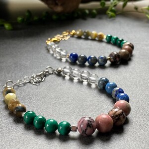 May include: Two beaded bracelets with a variety of colorful stones, including green, blue, pink, and brown. The bracelets are made with clear quartz, malachite, lapis lazuli, and rose quartz.