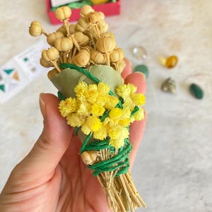 May include: A small bundle of dried flowers and herbs tied with green twine. The bundle is made up of yellow flowers, brown seed pods, and green leaves.