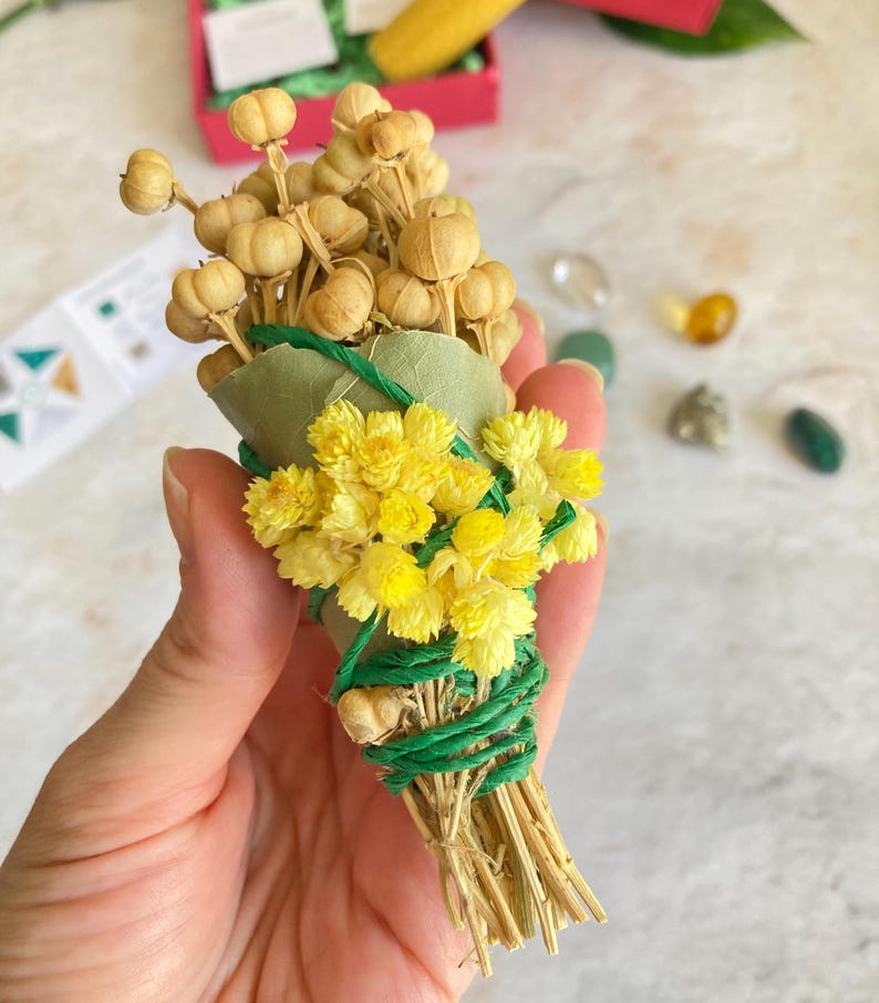 May include: A small bundle of dried flowers and herbs tied with green twine. The bundle is made up of yellow flowers, brown seed pods, and green leaves.