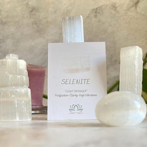May include: A collection of selenite crystals, including a tower, a sphere, and two sticks. The crystals are white and translucent. A card with the text "Selenite 'Light Bringer' Purification-Clarity-High Vibrations" is in the background.