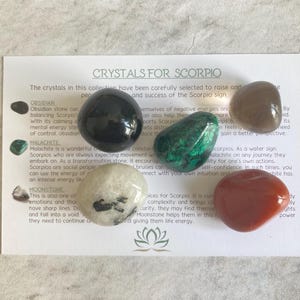 May include: A set of six crystals for Scorpio, including obsidian, malachite, moonstone, clear quartz, smoky quartz, and carnelian. The crystals are arranged on a white card with text describing the properties of each crystal.