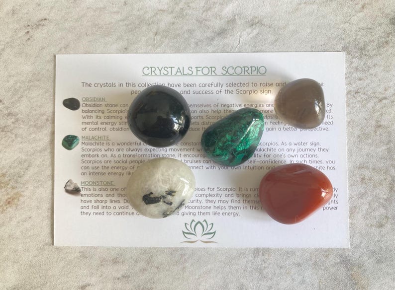May include: A set of six crystals for Scorpio, including obsidian, malachite, moonstone, clear quartz, smoky quartz, and carnelian. The crystals are arranged on a white card with text describing the properties of each crystal.
