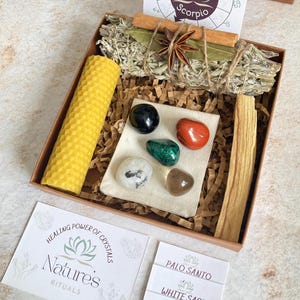 May include: A wooden box filled with items for a spiritual ritual. The box contains a white sage smudge stick, palo santo stick, beeswax candle, and a small pouch with crystals. The box also includes a card with the text "Healing Power of Crystals, Nature's Rituals".