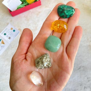 May include: A collection of five tumbled stones, including a clear quartz, a green aventurine, a green malachite, a gold pyrite, and a yellow citrine.