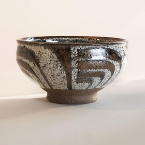 Puede incluir: Un cuenco de cerámica con exterior marrón oscuro y diseño en espiral blanco y marrón. El cuenco tiene forma redondeada con borde ligeramente ensanchado y base circular pequeña. El cuenco mide unos 15 cm de diámetro y 10 cm de alto.