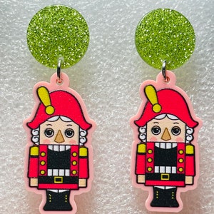 May include: A pair of dangle earrings featuring a pink nutcracker figurine with a red hat and a green glitter circle at the top.