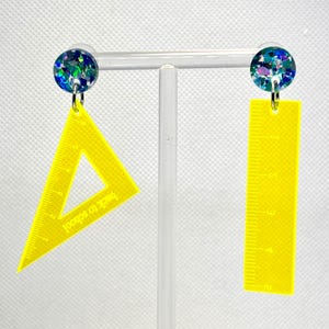 May include: A pair of novelty earrings with a glittery blue and green stud. One earring is a yellow triangle ruler, the other a yellow ruler. The text "back to school" is visible on the triangle ruler.