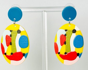 May include: A pair of colorful abstract oval earrings with a yellow background, red, blue, green, white, and black accents. The earrings are attached to a blue round disc.